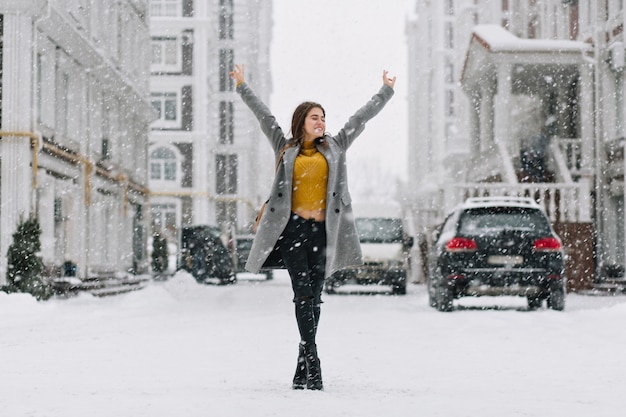 Free photo relaxing caucasian woman posing with hands up under snowfall on urban street. outdoor full-length photo of pretty woman in yellow sweater and gray coat enjoying weekend in winter town.