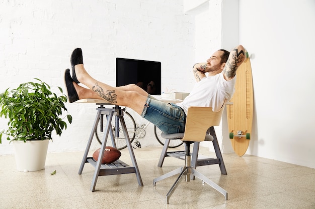 Relaxing tattoed freelancer dreamily looking in window while taking break from work, with his legs on table inside big white loft