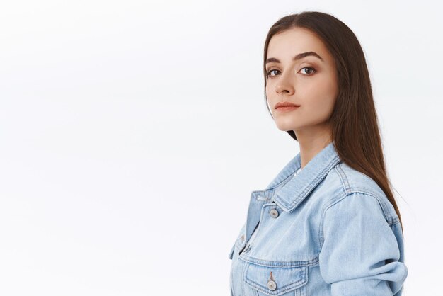 Reliable and determined goodlooking confident young sassy woman with long straight hair in denim jacket standing in profile and turn to camera with knowing look white background