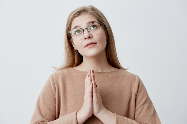 Religious girl with blonde hair in stylish eyewear pressings palms together and looking upwards, praying to God, begging for forgiveness or asking to make her dream come true. Emotions and feelings