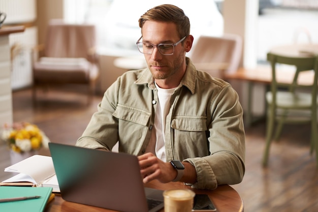 Free photo remote work and freelance concept handsome man in glasses sits with notebook and laptop studying