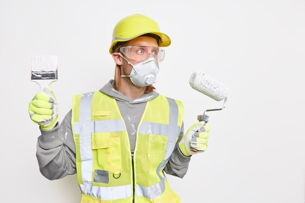 Renovation maintenance and engineering concept. Busy male labourer focused into distance with worried expression wears protective helmet safety gloves holds roller and brush follows repairing steps