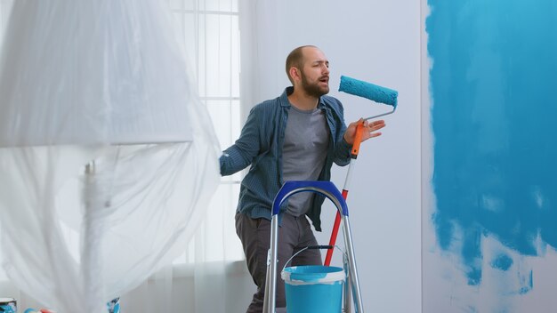 Repairman singing on roller brush dipped in blue paint while redecorating apartment. Housework, design, renovation. Home construction while renovating and improving. Repair and decorating.