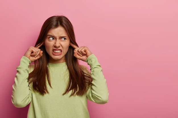 Free photo resentful annoyed woman plugs ears, irritated with loud noise, avoids annoying sound, dressed in casual green jumper, clenches teeth, frowns face, isolated on pink wall, blank space aside