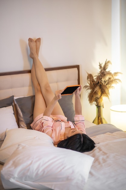 Rest. Young adult dark-haired woman in pink pajamas lying on her back with raised legs holding tablet above her looking at screen on bed in light room