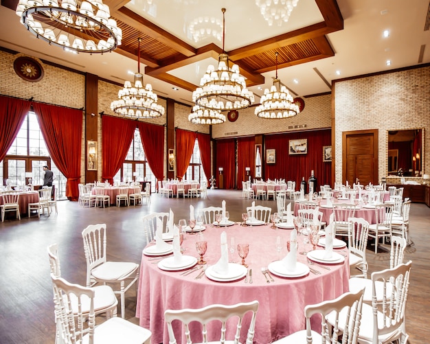 Restaurant hall with round tables, white napoleon chairs red curtains brick walls and chandeliers