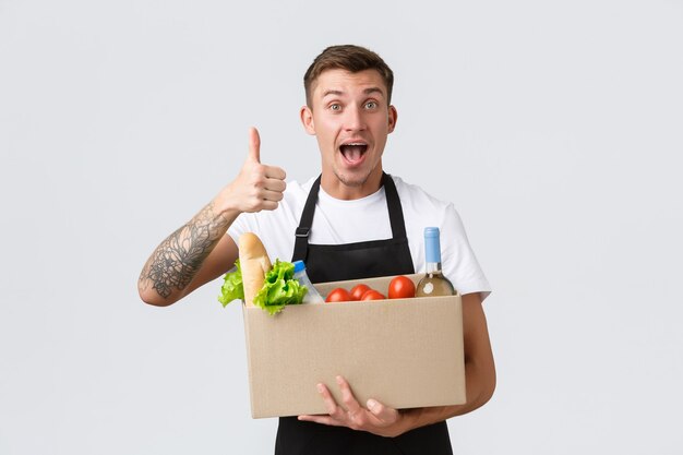 Retail grocery shopping and delivery concept handsome courier in black apron handing over box with g...