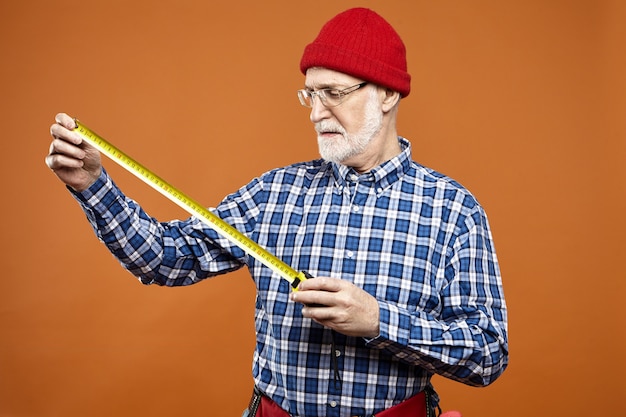 Free photo retired caucasian handyman or laborer wearing eyeglasses, red knitted hat and plaid shirt holding measuring tape while doing renovation, taking measurements, having serious concentrated look