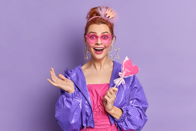 Free photo retro fashion model dressed in nineties style has happy expression nostalgia holds delicious candy poses over vivid purple background. fashion trends. redhead girl in trendy sunglasses purple jacket