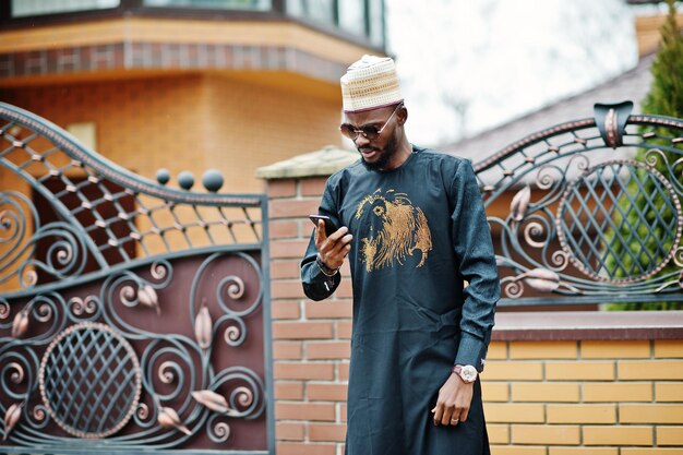 Rich african man in stylish traditional clothes and hat posed outdoor background his mansion speaking on mobile phone