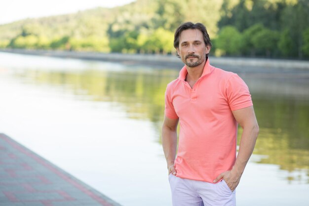 Rich man posing for photographer with his arms in pockets isolated on river. Middle-aged man in coral T-shirt owing huge factory.