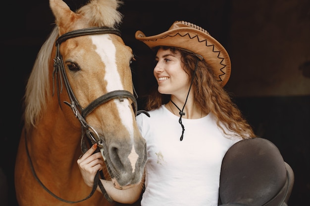 Rider woman holding a saddle in a stable. Woman has long hair and white t-shirt