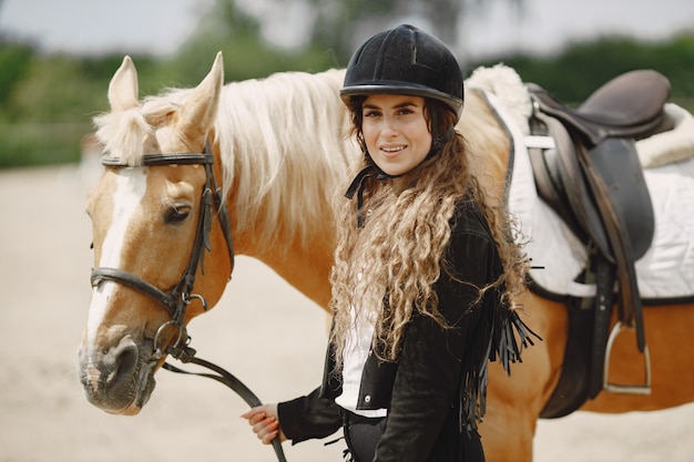 Rider woman looking at the camera. Woman has long hair and black clothes. Female equestrian touching a horse reins.