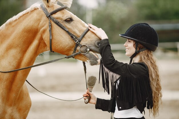 Rider woman talking to her horse on a ranch. Woman has long hair and black clothes. Female equestrian touching her brown horse.