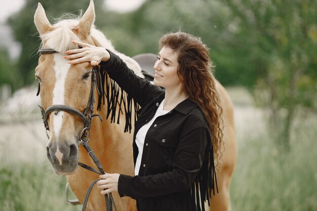 Rider woman talking to her horse on a ranch. Woman has long hair and black clothes. Female equestrian touching a horse reins.