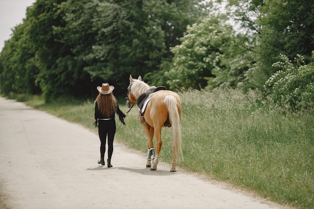 Rider woman walking with her horse on a ranch. Woman has long hair and black clothes. Female equestrian holding a horse reins.