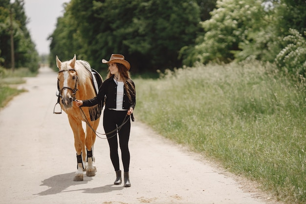 Free photo rider woman walking with her horse on a road. woman has long hair and black clothes. female equestrian holding a horse reins.