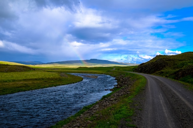 Free photo river in the middle of a road and grassy field with a rainbow in the distance at daytime