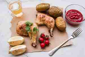 Free photo roasted chicken leg; boiled potato; sauce; cherry tomato; lemon; fork and beer for dinner on weathered background