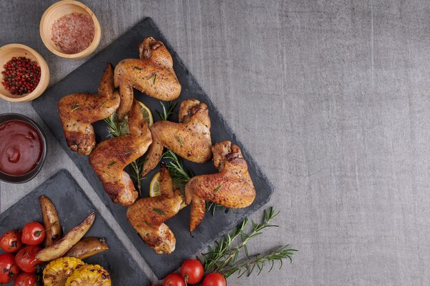 Free photo roasted chicken wings in barbecue sauce and mixed vegetable salad with pepper seeds rosemary, salt in black stone plate on gray stone table.