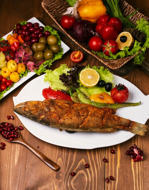 Roasted fried whole fish with grilled vegetables and lettuce. in white plate decorated with turshu on wooden table