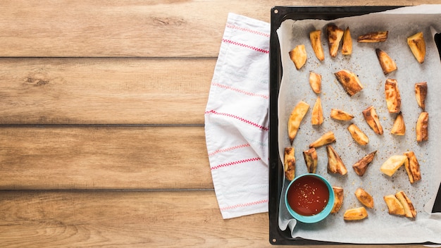 Free photo roasted potato pieces with ketchup in tray over wooden table