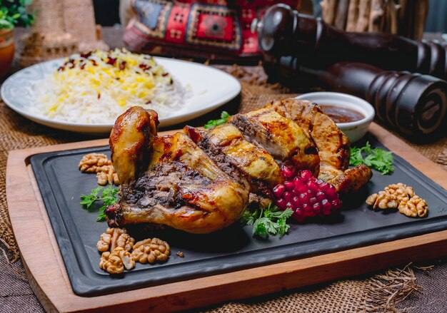 Roasted stuffed chicken decorated with pomegranate and walnuts on a black board and rice in a white plate on a wooden table