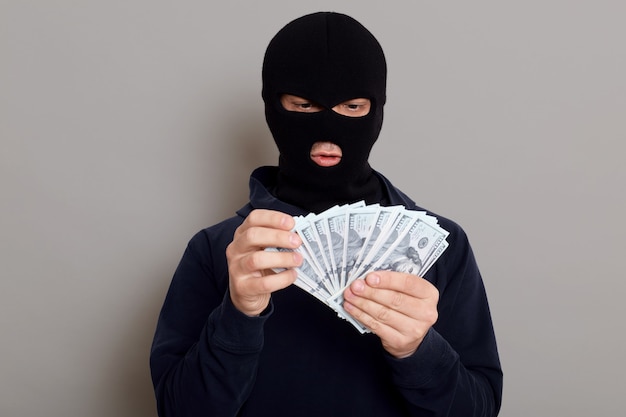 Robber man dressed in black hoodie stands with disguised face and holds a lot of money in his hands