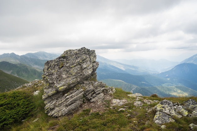 Rock on a hill covered in greenery with rocky mountains under a cloudy sky