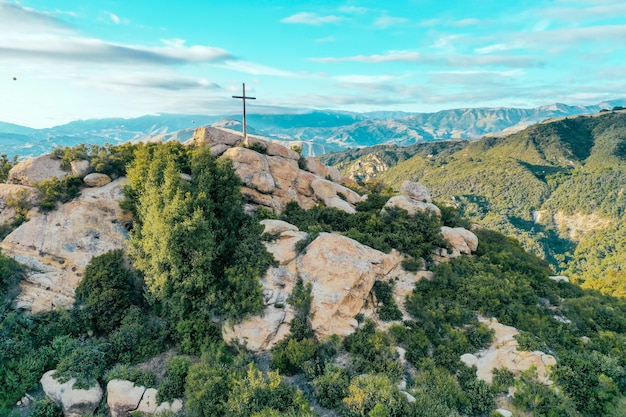 Rocky cliff covered in greenery with a cross put on the top and beautiful mountains