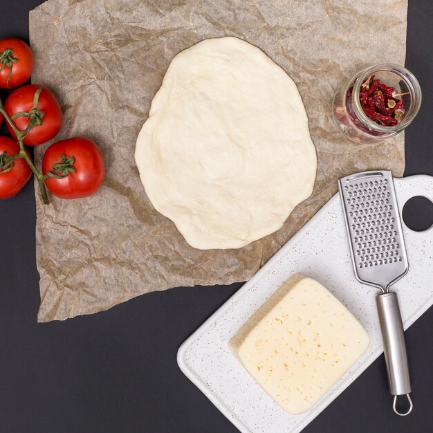 Rolled out pizza dough; tomatoes; cheese and dried red chili with kitchen utensil on black backdrop