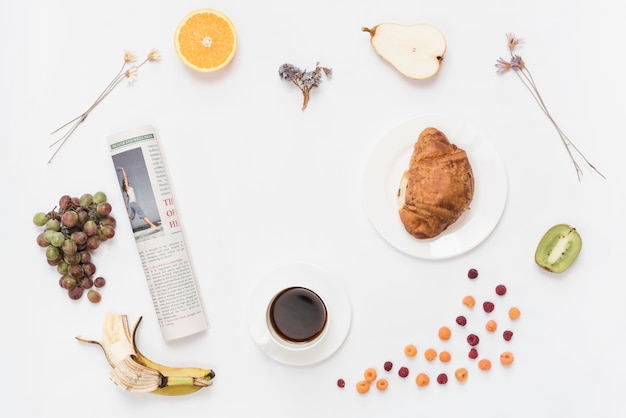 Free photo rolled up newspaper with coffee cup; croissant and fruits on white background