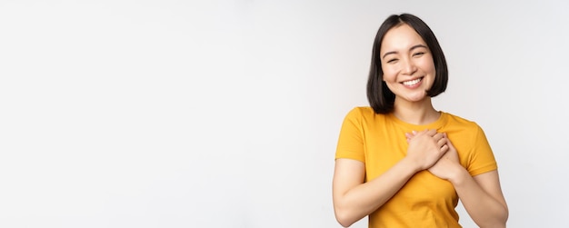 Romantic asian girfriend holding hands on heart smiling with care and tenderness standing in yellow