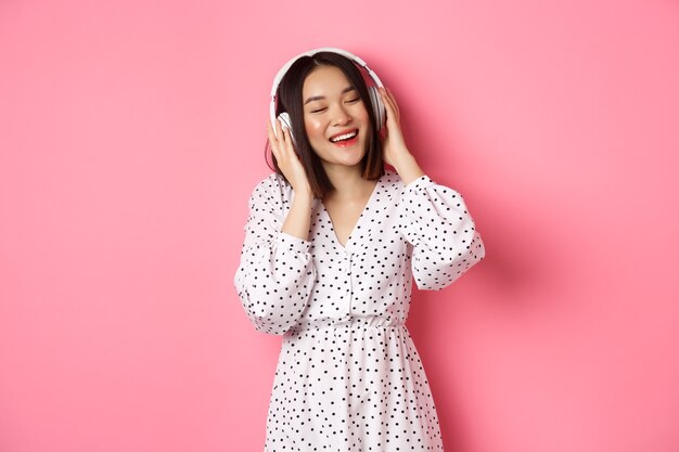 Romantic asian woman smiling happy, listening music in headphones and dancing, standing in trendy dress over pink background