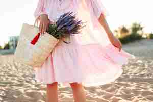 Free photo romantic blonde  female in cute pink  dress dancing and having fu on  the beach. holding straw bag and bouquet of lavender . freedom and nature concept.