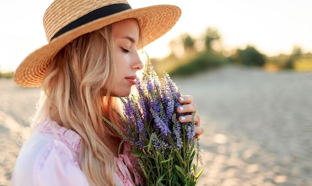 Free Photo romantic close up portrait  o charming blonde girl in straw hat  smells    flowers   on   the evening beach,  warm sunset colors. bouquet of lavender. details.