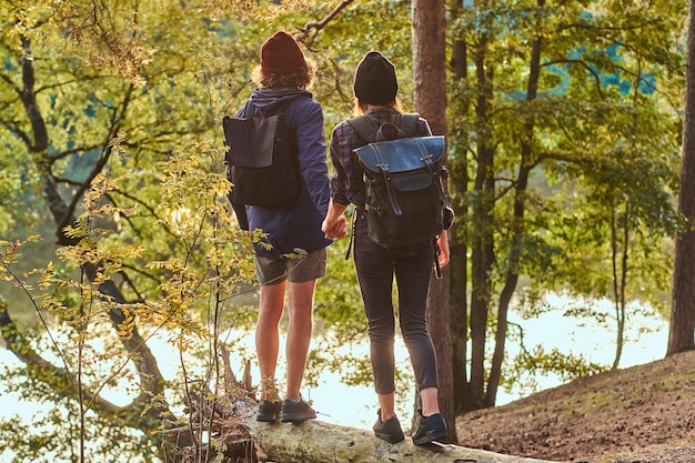 Free photo romantic couple is standing near river in green bright forest and looking foward. they have backpacks and hats.