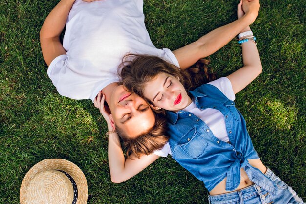 Romantic couple of young people lying on grass in park. They lay on the shoulders of each other and hold hands together. They keep eyes closed and look relaxed. View from above.