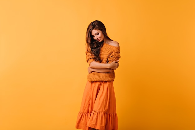 Free photo romantic dark-haired lady in cozy sweater posing on yellow. good-humoured female model in orange skirt looking down.