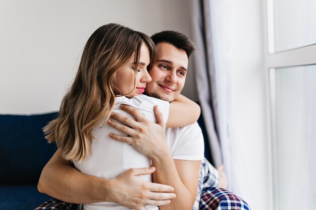 Romantic dark-haired man embracing his wife. Indoor shot of happy blonde girl relaxing on cozy couch.