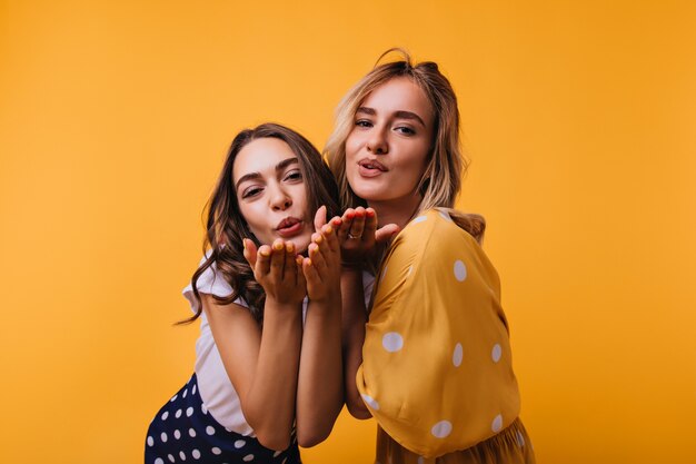 Romantic female friends in trendy clothes standing on yellow. Carefree girls sending air kisses.