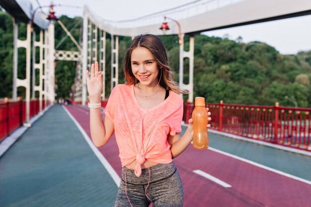 Romantic girl in pink t-short having fun on stadium. Lovable young woman with bottle of water laughing at cinder track.