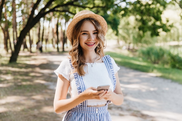 Romantic girl wears hat and white t-shirt smiling on nature. Adorable fair-haired woman enjoying walk in park.