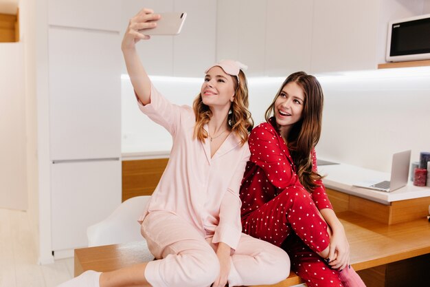 Romantic girls sitting on wooden table together and taking picture of themselves. Indoor shot of lovely ladies in pajamas making selfie in kitchen.