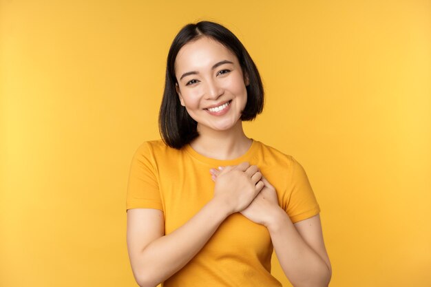 Romantic korean girl asian woman holding hands on heart smiling with care and tenderness standing over yellow background