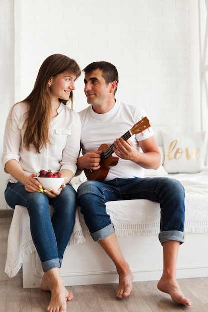 Romantic man playing ukulele and looking at his wife holding bowl of strawberries