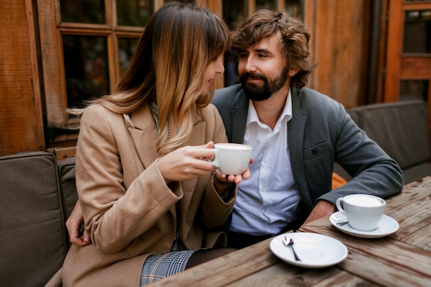 Romantic pensive woman with long wavy hairs hugging her husband with beard. Elegant couple sitting in cafe with hot cappuccino.