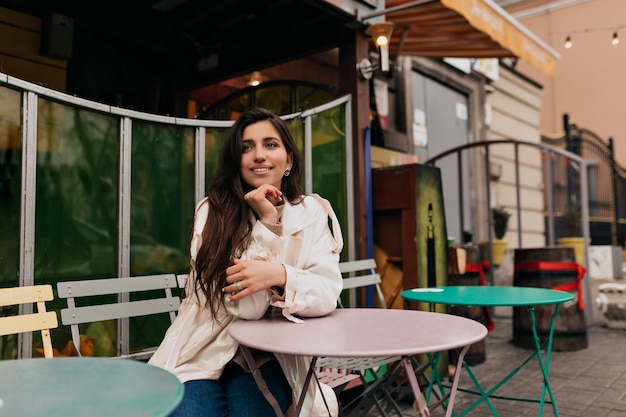 Free photo romantic shy girl with long hair wearing white coat sitting on french cafe outdoors and waiting for date