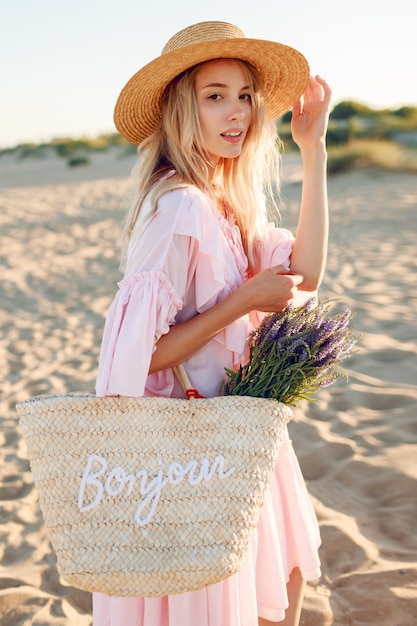 Romantic white woman in  trendy hat and  elegant pink dress posing on the beach.Holding straw bag  and bouquet of   flowers.