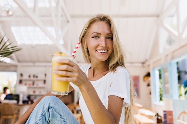 Free photo romantic woman in blue jeans drinking orange cocktail with pleasure. indoor shot of smiling blonde girl holding glass of cold juice while sitting in cafeteria.